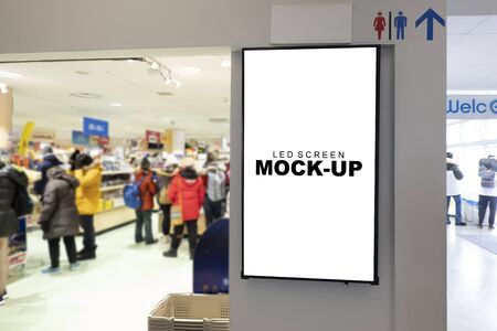 Mock up blank vertical LED board with clipping path in black frame on wall of shopping mall, blurred people wearing winter coat, empty space for insert advertising or announcement informationの写真素材