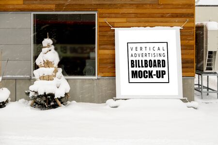 Mock up vertical blank outdoors signboard with clipping path hanging on panel of shop, moving snow cover on Christmas tree and walkway, empty space for insert advertising or announcement informationの写真素材