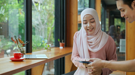 Selective focus, Young beautiful muslim woman wearing hijab use  laptop to working and Waiter come, She sign on paper slip for payment in coffee shopの写真素材
