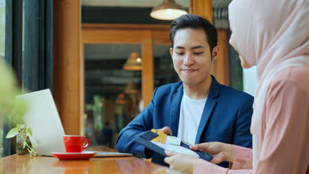 selective focus, Young muslim man working on laptop and Waitress wearing hijab come in with credit card payment slip for him sign, copy spaceの写真素材