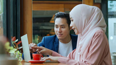 Selective focus, Young muslim man and beautiful woman wearing hijab working on laptop computer, They are point and look on screen and  talking together in coffee shop, muslim business working conceptの写真素材