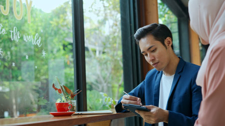 selective focus, Young muslim man working on laptop and Waitress wearing hijab come in with payment slip, He sign and pick up his credit cardの写真素材