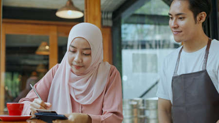Close up, Young beautiful muslim woman wearing hijab use  laptop to working and Waiter come, She sign on paper slip of credit card for payment in coffee shopの写真素材