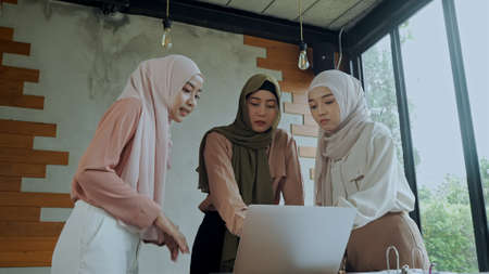 Young beautiful muslim women wearing hijab and use laptop while brainstorm meeting, they looking and point to monitor and talking together in meeting roomの写真素材