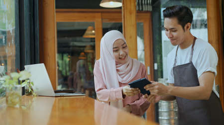 Young beautiful muslim woman wearing hijab sitting with laptop and Waiter come, she use credit card for payment in coffee shop, copy spaceの写真素材