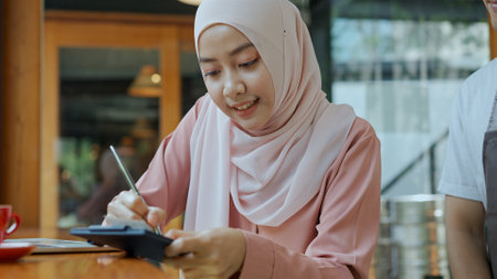 Selective focus, Young beautiful muslim woman wearing hijab use  laptop to working and Waiter come, She sign on paper slip of credit card for payment in coffee shopの写真素材