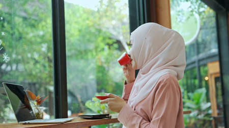 Side view, Young beautiful muslim woman wearing hijab sitting and drink hot coffee in red cup at coffee shop while use laptop computer working out of the office, copy spaceの写真素材