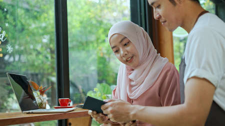 Selective focus, Young beautiful muslim woman wearing hijab typing on laptop, Waiter come in and She use credit card to payment in coffee shopの写真素材