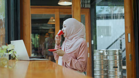 Young beautiful muslim woman wearing hijab sitting and drink hot coffee in red cup at coffee shop while use laptop computer working out of the office, copy spaceの写真素材