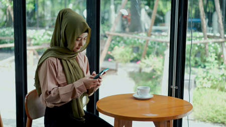 Young beautiful muslim woman sitting near glass window using smartphone chatting in coffee shop with happinessの写真素材