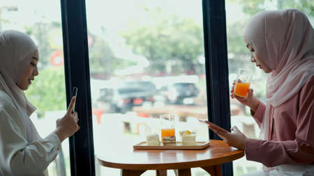 Cheerful young pretty muslim woman wearing hijab sitting near glass window in coffee shop with her friend, they are use smartphone to take photo with happinessの写真素材