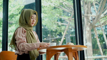 Young beautiful muslim woman wearing hijab sitting near glass window in coffee shop with bakery and coffee cupの写真素材