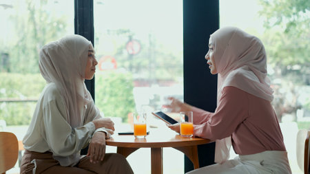 Young beautiful muslim woman and friend wearing hijab are sitting near glass window and used smartphone during talking together in coffee shop, muslim business working conceptの写真素材