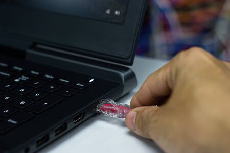 Closeup hand man connection red cable network connection to a Lan port of a  laptop on white table,...
