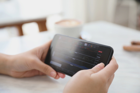 Female hand with smartphone trading stock online in coffee shop , Business conceptの写真素材