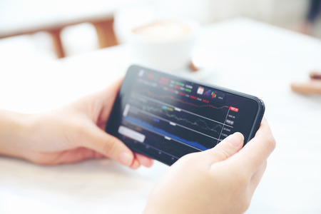 Female hand with smartphone trading stock online in coffee shop , Business concept