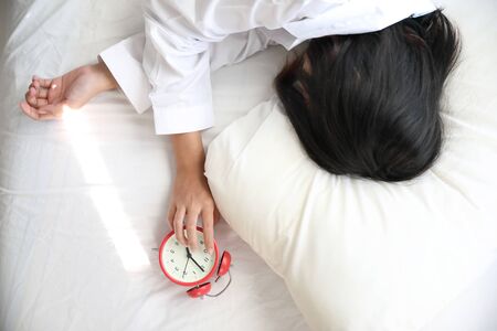 Asian woman lazy sleep waking up on bed with clock in white bedroomの写真素材