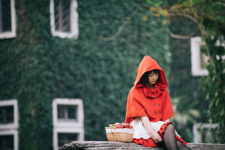 Portrait young woman with Little Red Riding Hood costume with apple and bread on basket sitting in green tree park backgroundの写真素材