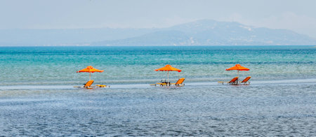 Three orange coloured sun beds and umbrellas left on the beach and the tide has come in and they are stranded in the waterの写真素材