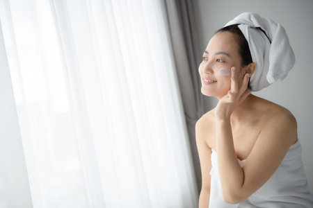 Happy young Asian woman applying face lotions while wearing a towel and touching her face.の写真素材