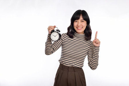 Smiling cheerful attractive young asian woman wearing sweater shirt holding alarm clock, point finger above and looking camera isolated on white background, studio portrait.の写真素材