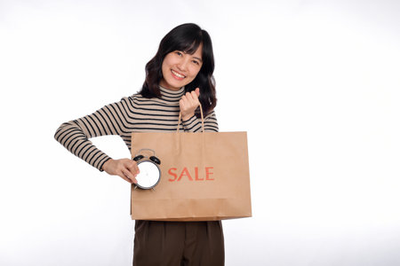 Portrait of happy young Asian woman with sweater shirt holding alarm clock and shopping paper back isolated on white background.の写真素材
