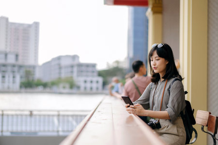 Young Asian woman backpack traveler using mobile phone in express boat pier on Chao Phraya River in Bangkok. Traveler checking out side streets.の写真素材