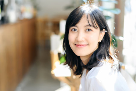 Brightly smiling beautiful young asian woman sitting in a coffee cafe in sunny dayの写真素材