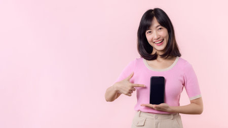 Portrait beautiful young asian woman happy smile showing smartphone screen with pointing finger hand gesture to mobile phone isolated on pink pastel studio background.の写真素材