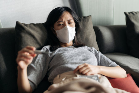 Sick asian woman reading clinical thermometer while cough on a sofa bed.の写真素材