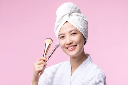Portrait young happy asian woman with natural make up face holding cosmetic skin powder blusher isolated on pink background. Female apply skincare brush treatment. beauty product, cosmetology concept.の写真素材