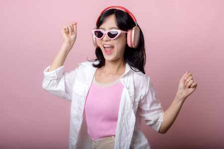 young happy asian woman model with stylish trendy sun glasses enjoy listening music by headphone audio and dancing isolated on pink studio background. technology, girl fashion, accessory concept.の写真素材