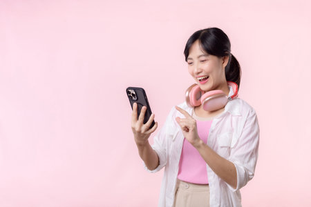 Portrait attractive young asian woman happy smile using smartphone with earphone, headphone isolated on pink studio background. Pretty female person using mobile phone. Music online lifestyle concept.の写真素材
