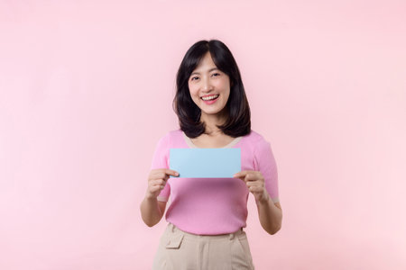 Portrait young happy woman model holding and showing blank space paper for advertisement information message poster with thumb up or point finger gesture isolated on pink pastel studio background.の写真素材