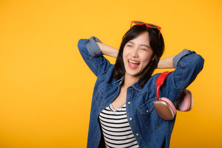 Fashion portrait asian young woman wearing denim clothes, red glasses and headphone isolated on yellow background. Lifestyle people concept.の写真素材