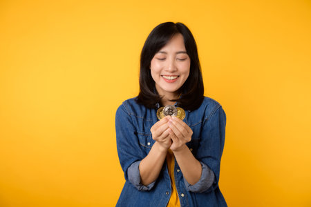 Happy asian young woman wearing yellow t-shirt denim shirt holding digital crypto currency coin isolated on yellow background. Digital currency financial concept.の写真素材