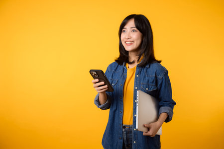 Portrait asian young woman wearing yellow t-shirt and denim shirt holding laptop and smartphone isolated on yellow studio background. Technology laptop advertise concept.の写真素材