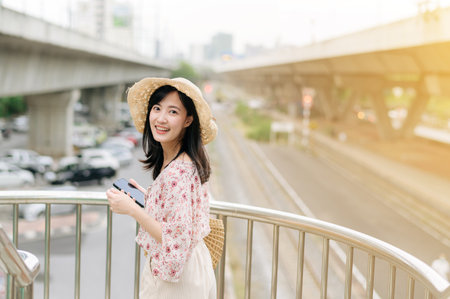 asian young woman traveler with weaving basket using mobile phone and standing on overpass with railway background. Journey trip lifestyle, world travel explorer or Asia summer tourism concept.の写真素材
