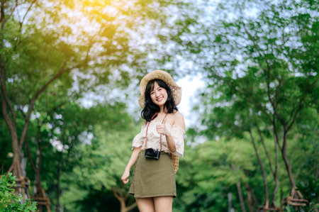 Portrait of young asian woman traveler with weaving hat and basket and a camera on green public park nature background. Journey trip lifestyle, world travel explorer or Asia summer tourism concept.の写真素材
