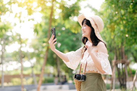Portrait of young asian woman traveler with weaving hat, basket, mobile phone and camera on green public park background. Journey trip lifestyle, world travel explorer or Asia summer tourism concept.の写真素材