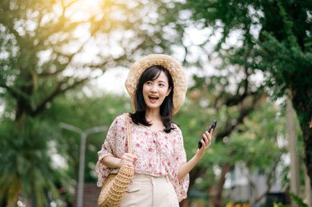 Portrait of young asian woman traveler with weaving hat and basket using mobile phone on green park nature background. Journey trip lifestyle, world travel explorer or Asia summer tourism concept.の写真素材