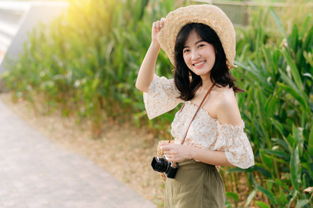 Portrait of young asian woman traveler with weaving hat and basket and a camera on green public park nature background. Journey trip lifestyle, world travel explorer or Asia summer tourism concept.の写真素材