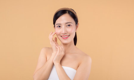 Beautiful asian girl model touching fresh glowing hydrated facial skin on beige background closeup. Beauty face young woman with natural makeup and healthy skin portrait. Skin care conceptの写真素材