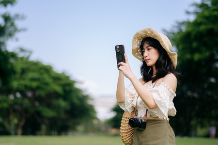 Portrait of young asian woman traveler with weaving hat, basket, mobile phone and camera on green public park background. Journey trip lifestyle, world travel explorer or Asia summer tourism concept.の写真素材