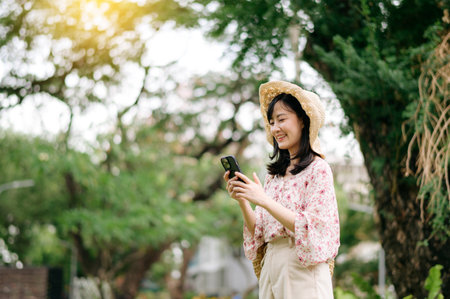 Portrait of young asian woman traveler with weaving hat and basket using mobile phone on green park nature background. Journey trip lifestyle, world travel explorer or Asia summer tourism concept.の写真素材