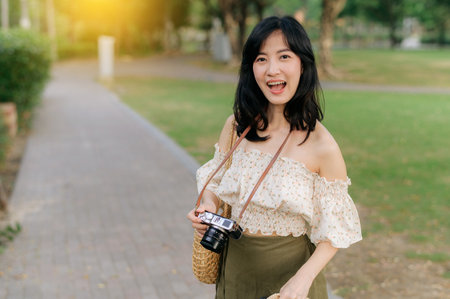 Portrait of young asian woman traveler with weaving hat and basket and a camera on green public park...