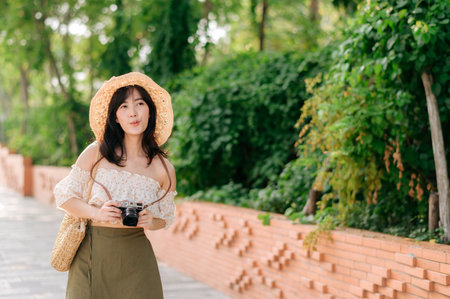 Portrait of young asian woman traveler with weaving hat and basket and a camera on green public park nature background. Journey trip lifestyle, world travel explorer or Asia summer tourism concept.の写真素材