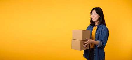 Portrait young asian woman wearing yellow t-shirt and denim shirt holding parcel box isolated on yellow studio background, Delivery courier and shipping service concept.の写真素材