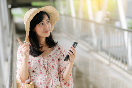 asian young woman traveler with weaving basket using mobile phone and standing on overpass. Journey trip lifestyle, world travel explorer or Asia summer tourism concept.の写真素材