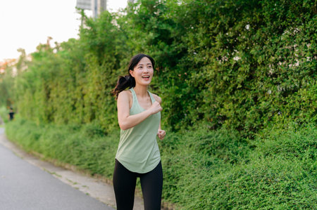 Fit young Asian woman jogging in park smiling happy running and enjoying a healthy outdoor lifestyle. Female jogger. Fitness runner girl in public park. healthy lifestyle and wellness being conceptの写真素材